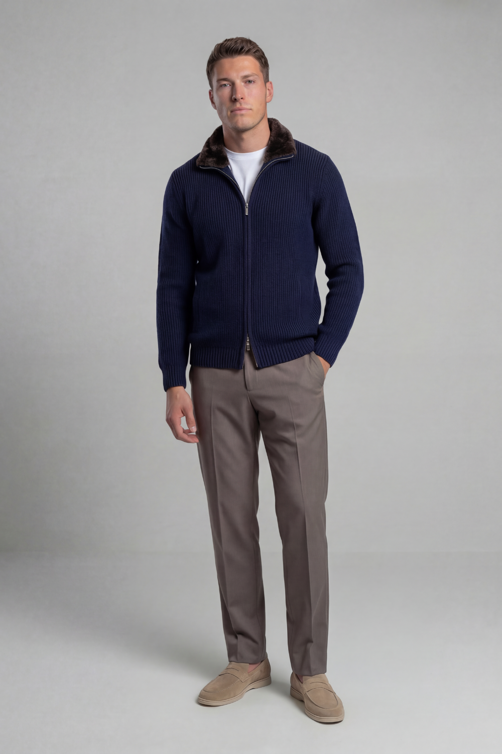 Man wearing a navy blue sweater with a fur collar, white shirt, and brown pants on a gray background